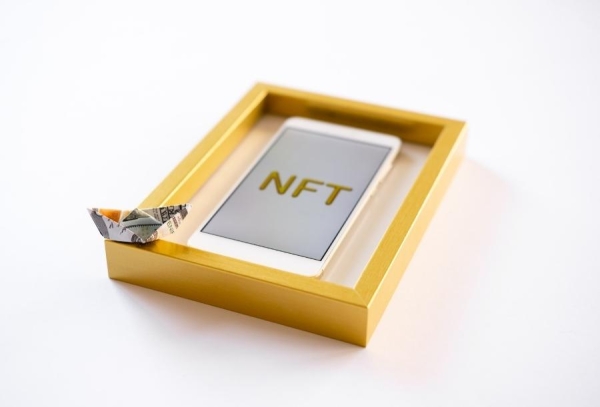 Papírová lodička z bankovky položená na zlatém rámu s telefonem a nápisem NFT